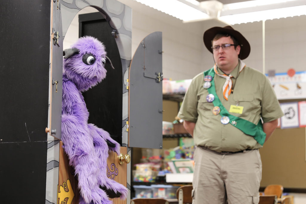 A man in a purple shirt and hat stands beside a purple stuffed animal.